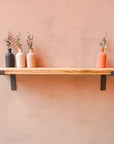 A reclaimed rustic wooden radiator shelf with propped metal brackets, displaying four bottles with flowers in shades of white, beige, black, and red.