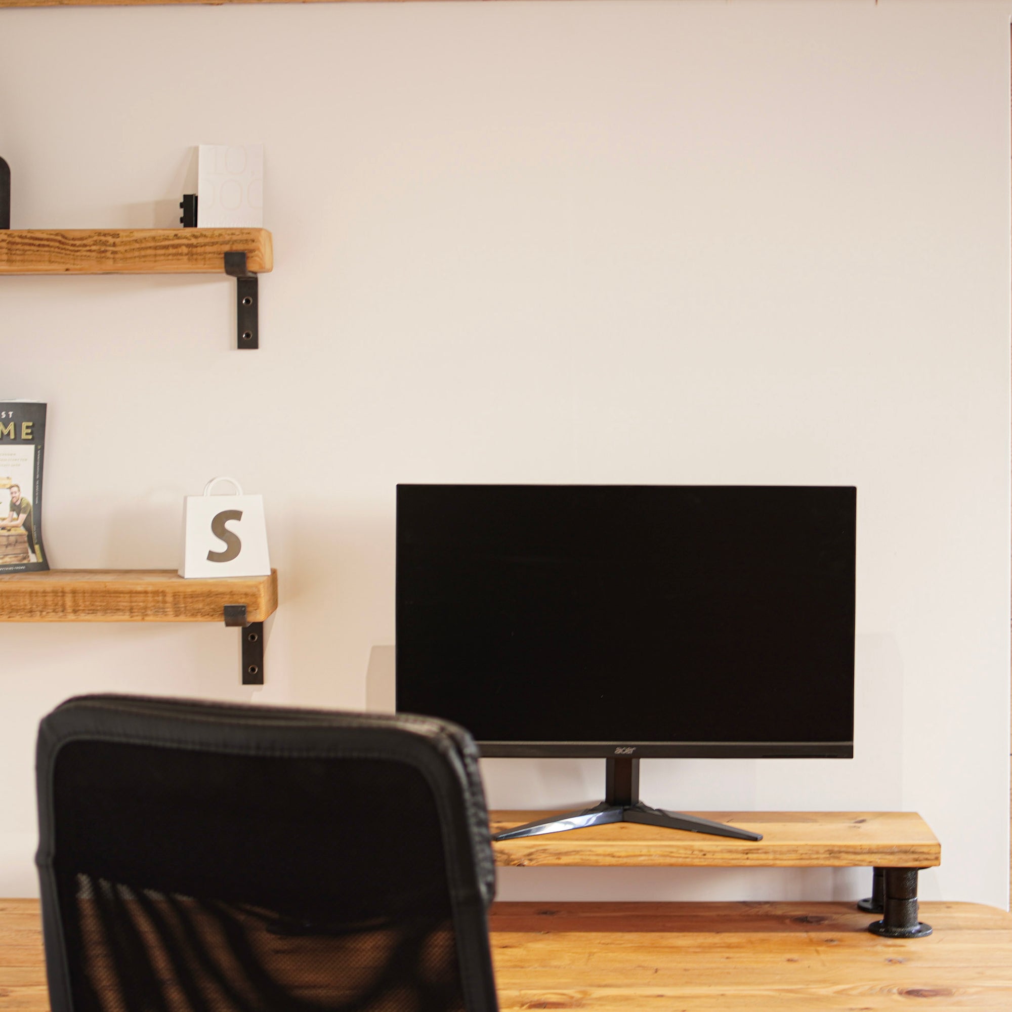 Monitor stand made from reclaimed scaffolding board, positioned on a wooden desk with a monitor and shelves in the background.