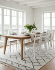 Dining room with wooden table and white chairs near large windows.