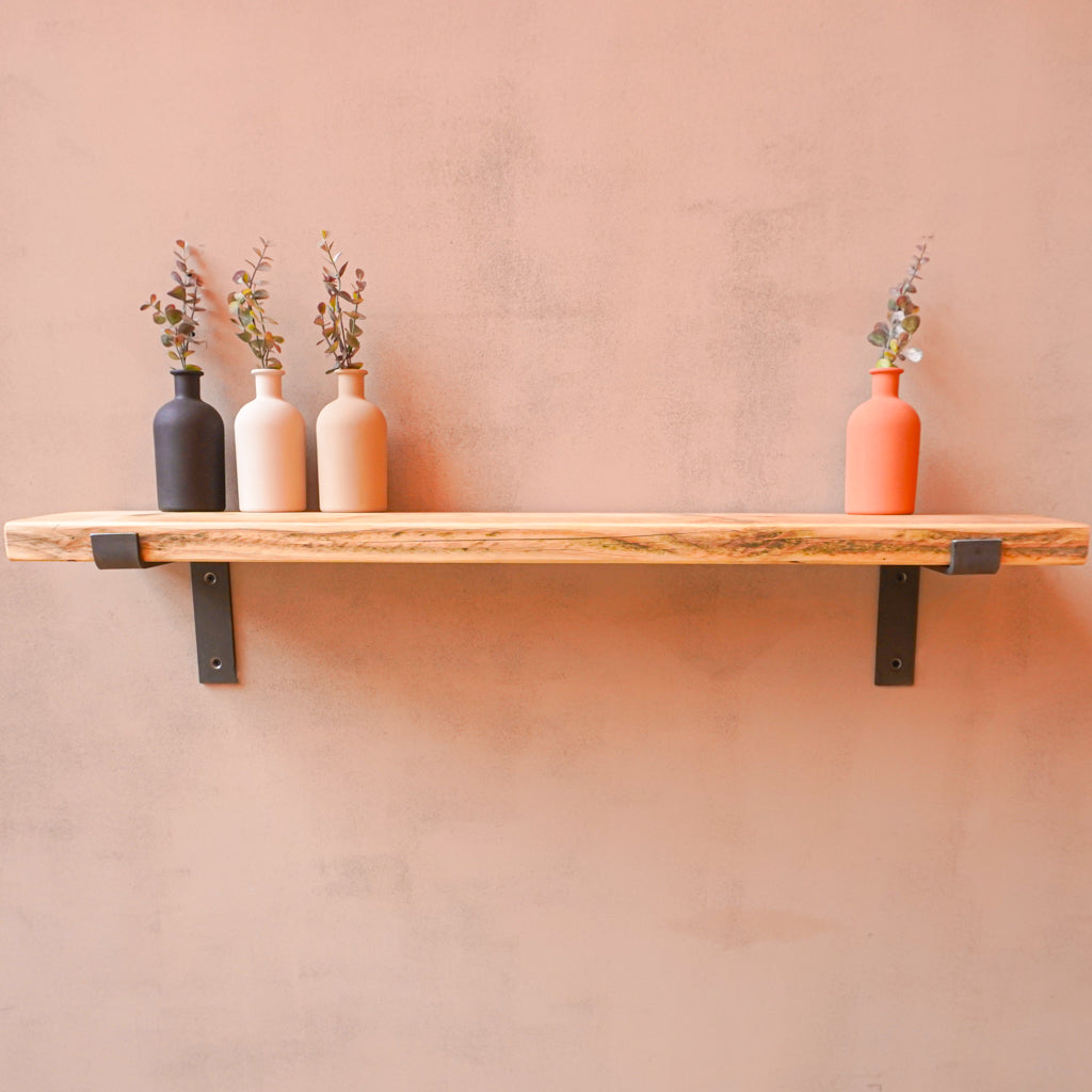 A reclaimed rustic wooden radiator shelf with propped metal brackets, displaying four bottles with flowers in shades of white, beige, black, and red.