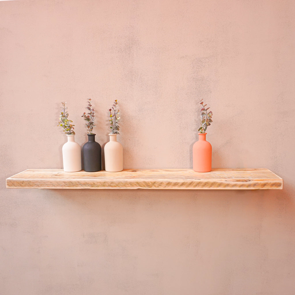 A rustic wooden shelf with floating brackets mounted on a wall, made from reclaimed scaffolding board displaying three vases containing flowers.