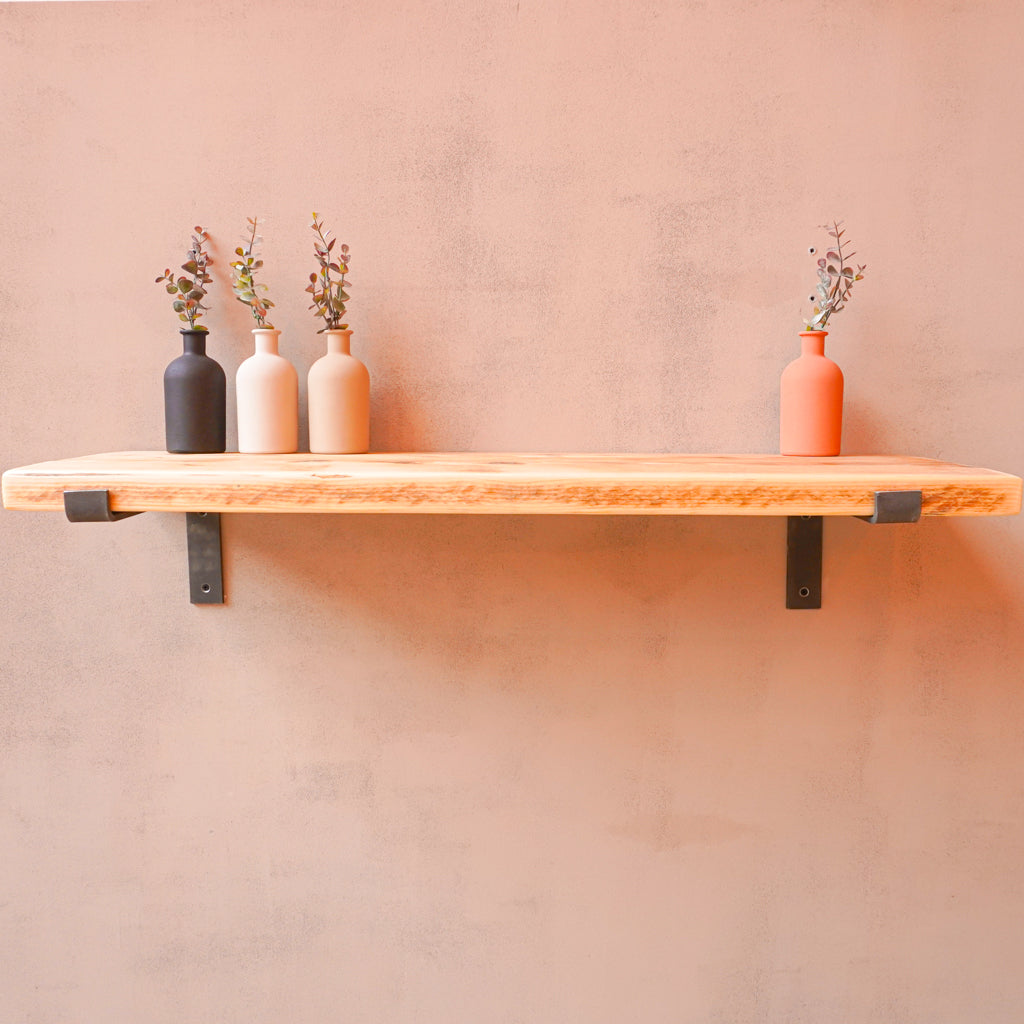 A reclaimed rustic wooden shelf mounted on a wall, using propped metal brackets, showcasing three vases.